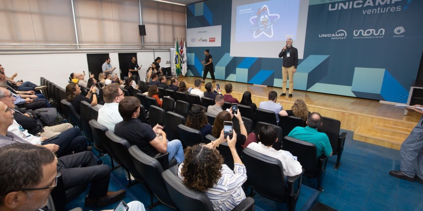 Foto colorida mostra auditório moderno com pessoas assistindo a uma apresentação no palco, onde um homem fala ao microfone. Fim da descrição.