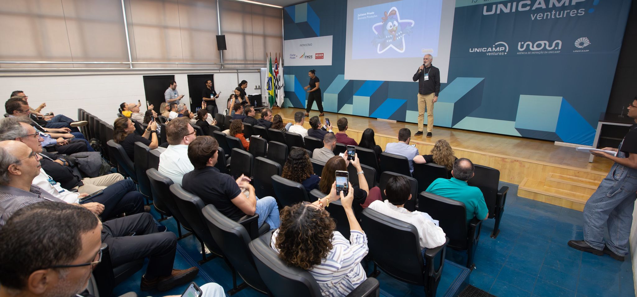 Foto colorida mostra auditório moderno com pessoas assistindo a uma apresentação no palco, onde um homem fala ao microfone. Fim da descrição.