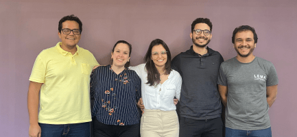 Foto de cinco pessoas adultas sorridentes posando juntos em frente a uma parede roxa. Eles estão em um ambiente interno. À esquerda, um homem usando uma camisa amarela está sorrindo e com o braço ao redor de uma mulher que usa uma camisa listrada de azul e com detalhes florais. No centro, uma mulher com uma blusa branca e óculos está sorrindo com o braço em volta de um homem de barba e óculos, que está usando uma camisa polo escura. À direita, há outro homem sorrindo, usando uma camiseta cinza com o logo "LEMA". Todos estão próximos uns dos outros e pousando para a foto. Fim de descrição.