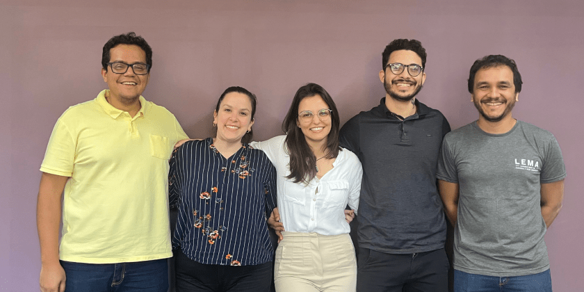 Foto de cinco pessoas adultas sorridentes posando juntos em frente a uma parede roxa. Eles estão em um ambiente interno. À esquerda, um homem usando uma camisa amarela está sorrindo e com o braço ao redor de uma mulher que usa uma camisa listrada de azul e com detalhes florais. No centro, uma mulher com uma blusa branca e óculos está sorrindo com o braço em volta de um homem de barba e óculos, que está usando uma camisa polo escura. À direita, há outro homem sorrindo, usando uma camiseta cinza com o logo "LEMA". Todos estão próximos uns dos outros e pousando para a foto. Fim de descrição.