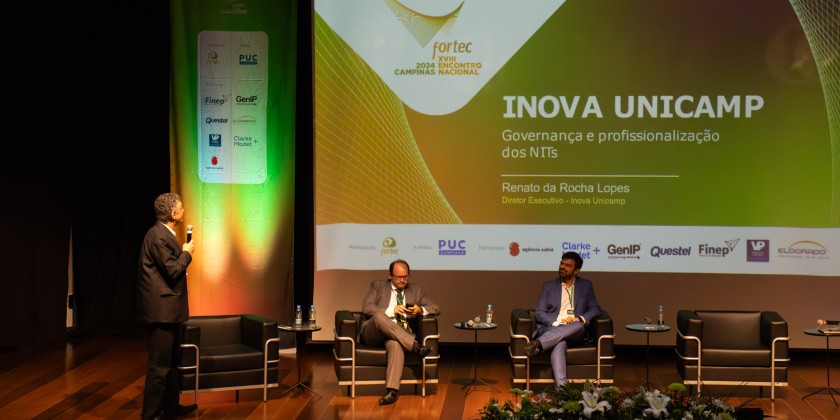 Fotografia colorida em ambiente interno. Professor Renato Lopes, um homem branco de terno completo, segura um microfone. Ele está de perfil, em cima do palco, virado para o telão, onde se vê um slide com o título "Inova Unicamp: Governança e profissionalização dos NITs. Renato da Rocha Lopes. Diretor Executivo - Inova Unicamp". No palco há quatro poltronas, duas estão ocupadas por outros dois participantes do painel.