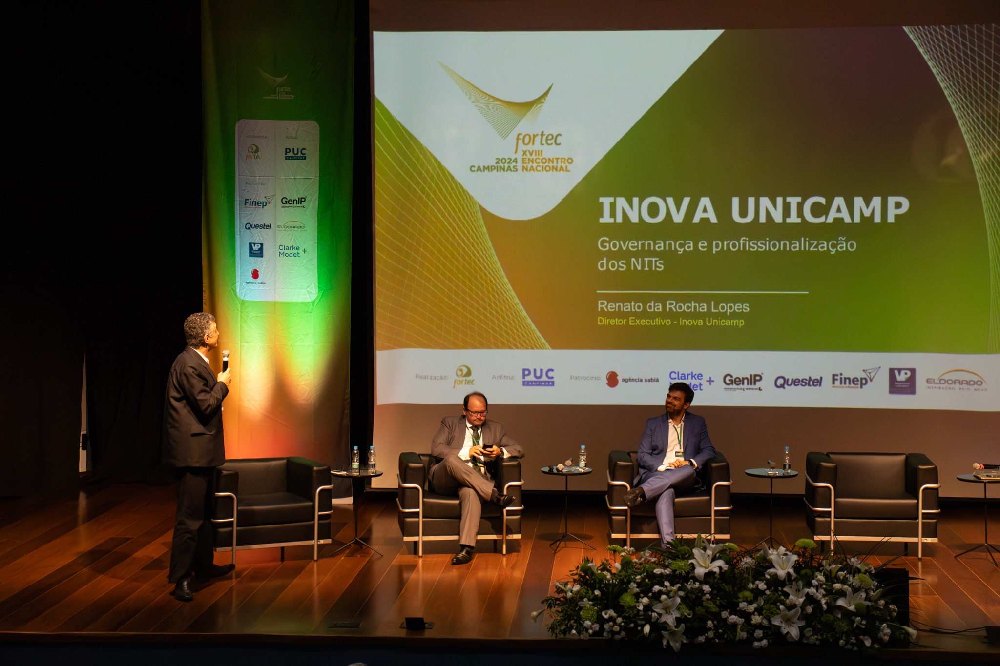 Fotografia colorida em ambiente interno. Professor Renato Lopes, um homem branco de terno completo, segura um microfone. Ele está de perfil, em cima do palco, virado para o telão, onde se vê um slide com o título "Inova Unicamp: Governança e profissionalização dos NITs. Renato da Rocha Lopes. Diretor Executivo - Inova Unicamp". No palco há quatro poltronas, duas estão ocupadas por outros dois participantes do painel.