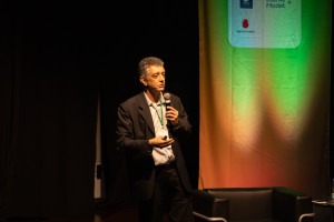 Fotografia colorida em ambiente interno. Professor Renato Lopes, um homem branco de terno completo, segura um microfone. Ele está de perfil, em cima do palco.