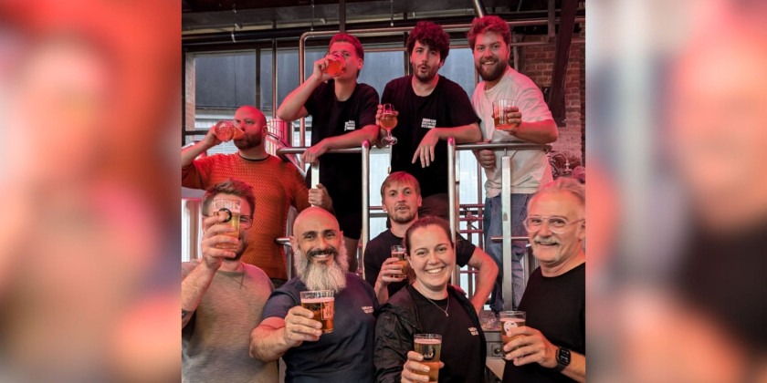 Foto colorida em espaço interno de cervejaria mostra nove pessoas sorindo e brindando com copos de cerveja. Fim da descrição.