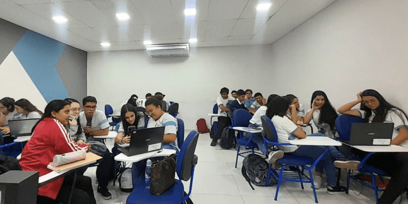 A imagem mostra uma sala de aula com mesas e cadeiras, em que os alunos estão dispostos por elas em grupos. Eles estão sentados e olhando para os computadores em cima das mesas. Os alunos estão participando da competição de empreendedorismo e concorrem para serem finalistas do Inova Jovem 2024. Fim da descrição.