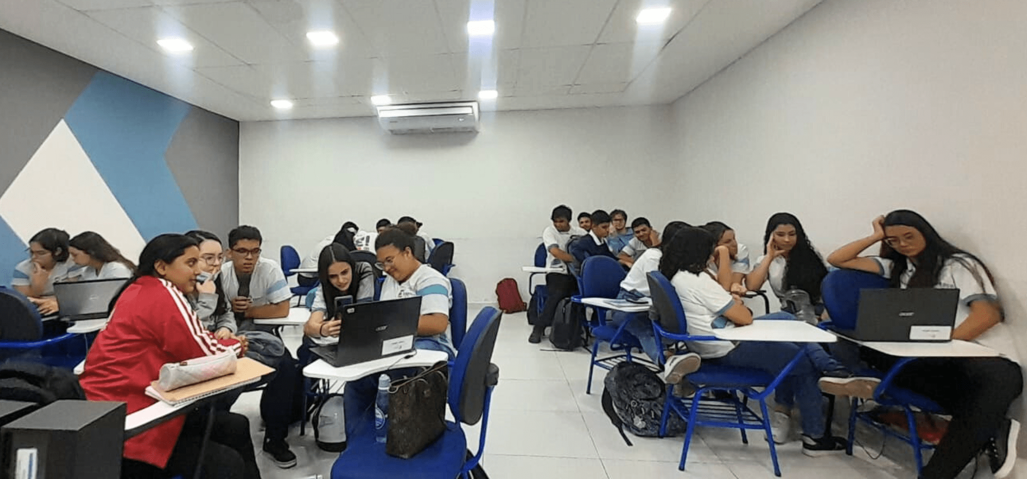 A imagem mostra uma sala de aula com mesas e cadeiras, em que os alunos estão dispostos por elas em grupos. Eles estão sentados e olhando para os computadores em cima das mesas. Os alunos estão participando da competição de empreendedorismo e concorrem para serem finalistas do Inova Jovem 2024. Fim da descrição.