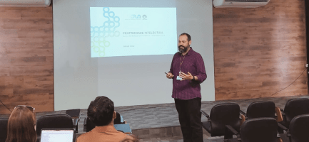 Fotografia. Imagem mostra um homem branco em pé, em frente a uma apresentação de slides com o título "Propriedade Intelectual". O registro também capta algumas pessoas sentadas na plateia, para onde o homem olha enquanto faz a apresentação. Fim da descrição.