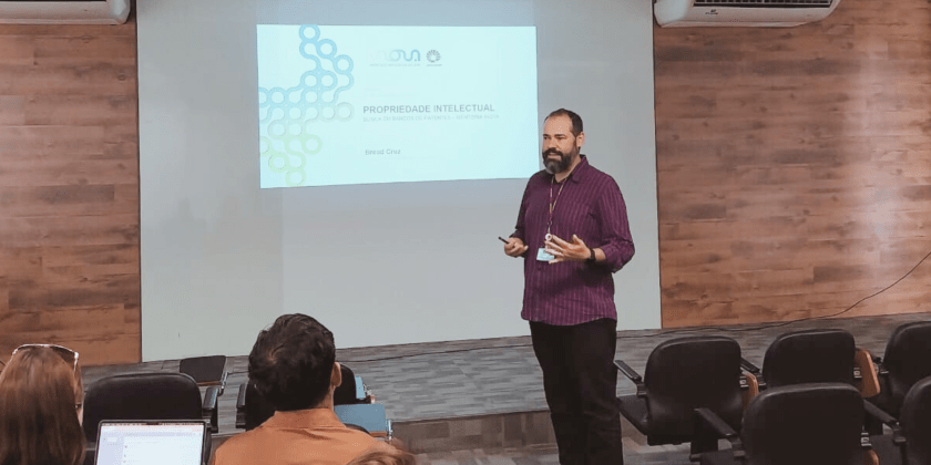 Fotografia. Imagem mostra um homem branco em pé, em frente a uma apresentação de slides com o título "Propriedade Intelectual". O registro também capta algumas pessoas sentadas na plateia, para onde o homem olha enquanto faz a apresentação. Fim da descrição.