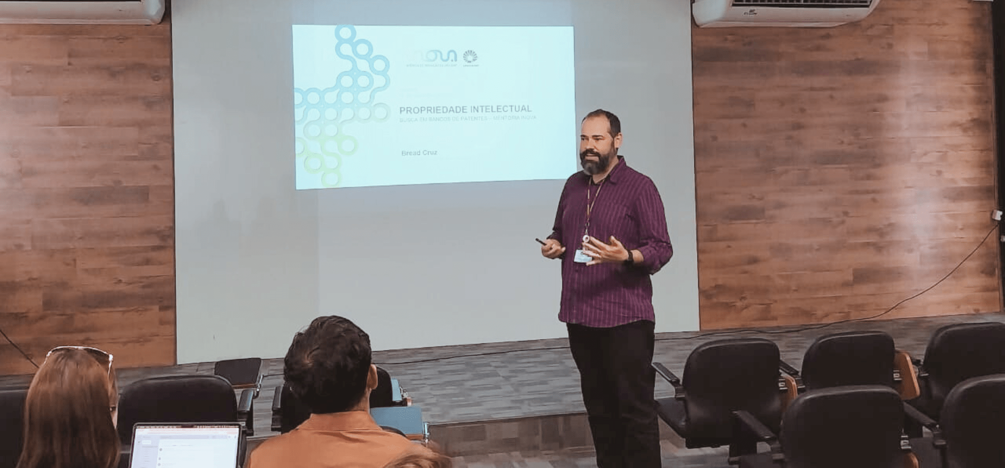 Fotografia. Imagem mostra um homem branco em pé, em frente a uma apresentação de slides com o título "Propriedade Intelectual". O registro também capta algumas pessoas sentadas na plateia, para onde o homem olha enquanto faz a apresentação. Fim da descrição.