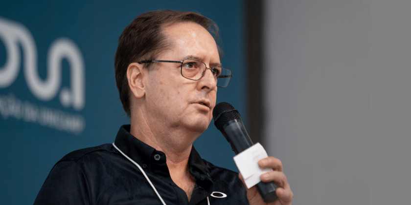 Fotografia colorida de um homem com uma microfone na mão direita. Ele está no palco fazendo sua apresentação. O fundo da fotografia está desfocado.