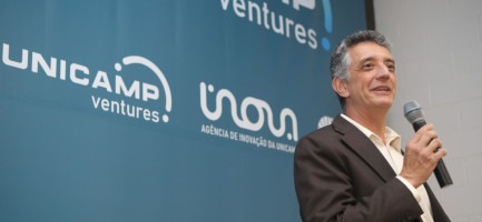 Foto colorida do professor Renato Lopes, diretor-executivo da Inova Unicamp, um homem branco de terno preto e camisa branca, que fala ao microfone. Ao fundo há um banner azul com os logotipos Unicamp Ventures, Inova e Unicamp. Fim da descrição.