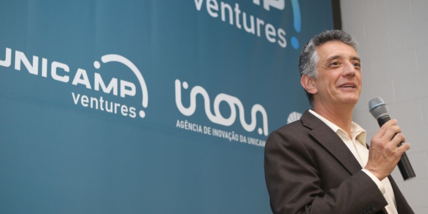 Foto colorida do professor Renato Lopes, diretor-executivo da Inova Unicamp, um homem branco de terno preto e camisa branca, que fala ao microfone. Ao fundo há um banner azul com os logotipos Unicamp Ventures, Inova e Unicamp. Fim da descrição.