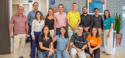 Foto posada de participantes do eMerge Americas e de colaboradores da Inova Unicamp. A imagem com treze pessoas que estão em pé ou agachadas, lado a lado. Elas estão olhando para a câmera, em ambiente interno com fundo de parede cinza e uma porta branca com vidros que dá acesso a outro local. Fim da descrição.