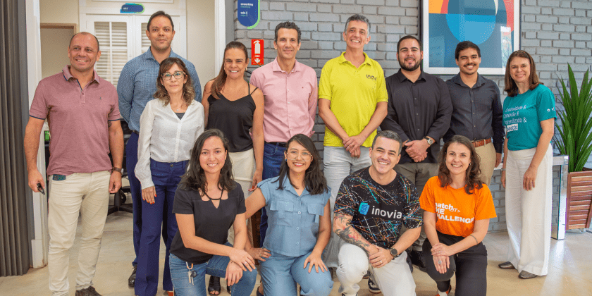 Foto posada de participantes do eMerge Americas e de colaboradores da Inova Unicamp. A imagem com treze pessoas que estão em pé ou agachadas, lado a lado. Elas estão olhando para a câmera, em ambiente interno com fundo de parede cinza e uma porta branca com vidros que dá acesso a outro local. Fim da descrição.