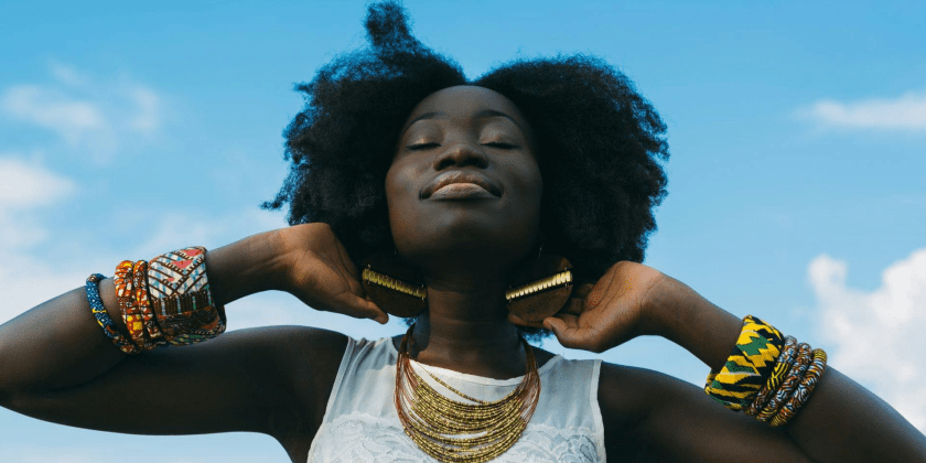 A imagem mostra uma mulher negra posando para a foto em um fundo azul, que mostra o céu e algumas nuvens. A mulher está segurando o cabelo com as duas mãos. Fim da descrição.