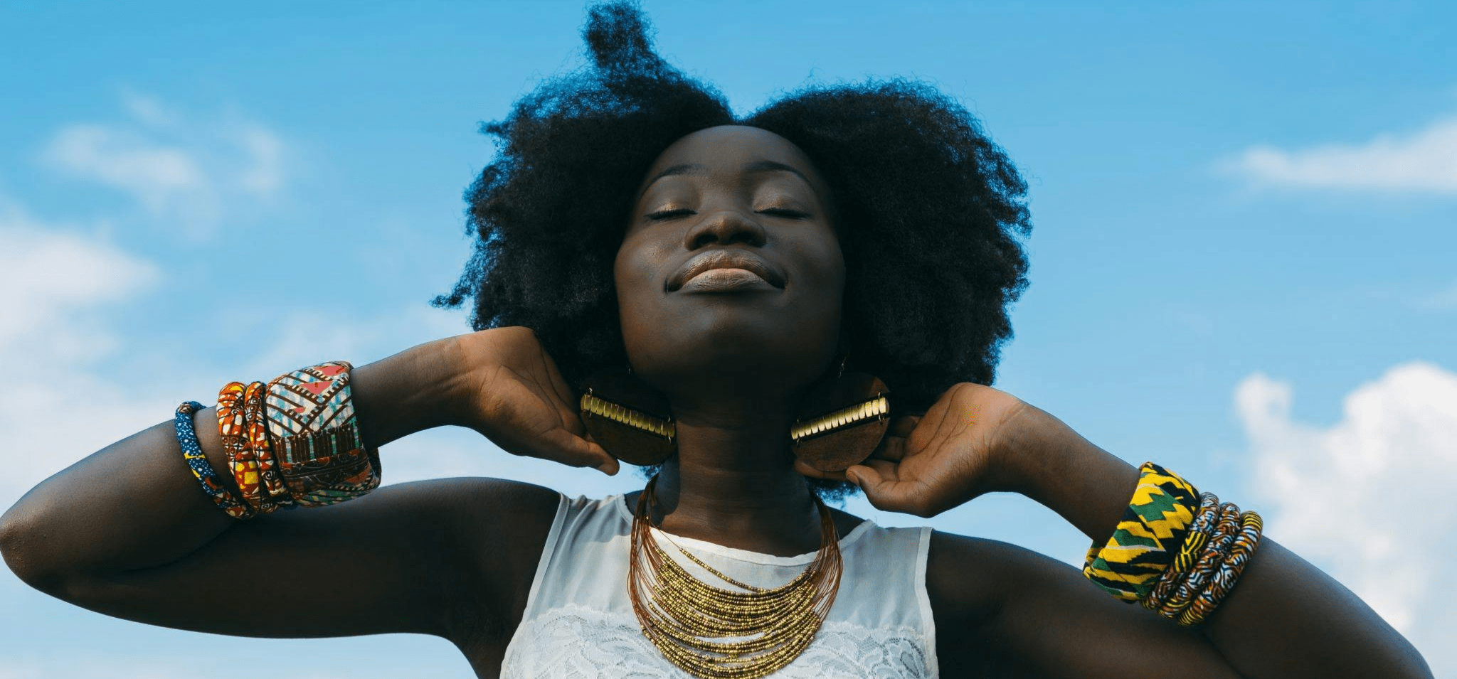 A imagem mostra uma mulher negra posando para a foto em um fundo azul, que mostra o céu e algumas nuvens. A mulher está segurando o cabelo com as duas mãos. Fim da descrição.