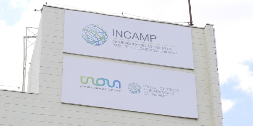 Placa com logo da Incamp no topo do prédio da Incubadora. Abaixo da placa da Incamp está uma placa com os logos da Inova Unicamp e do Parque Científico e Tecnológico da Unicamp Fim da descrição.