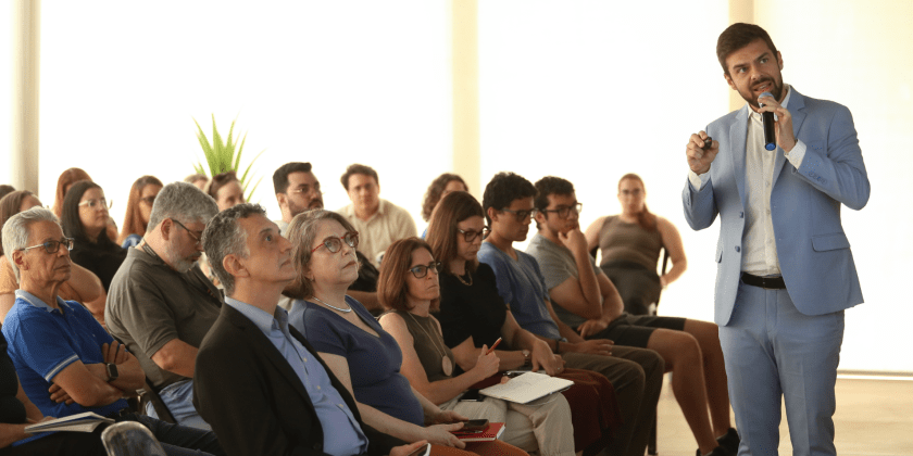 Uma fotografia colorida, em destaque no canto direiro está Rafael Carvalho de Fassio durante sua fala. Na esquerda, a platéia assiste atenta à apresentação. Fim da descrição.