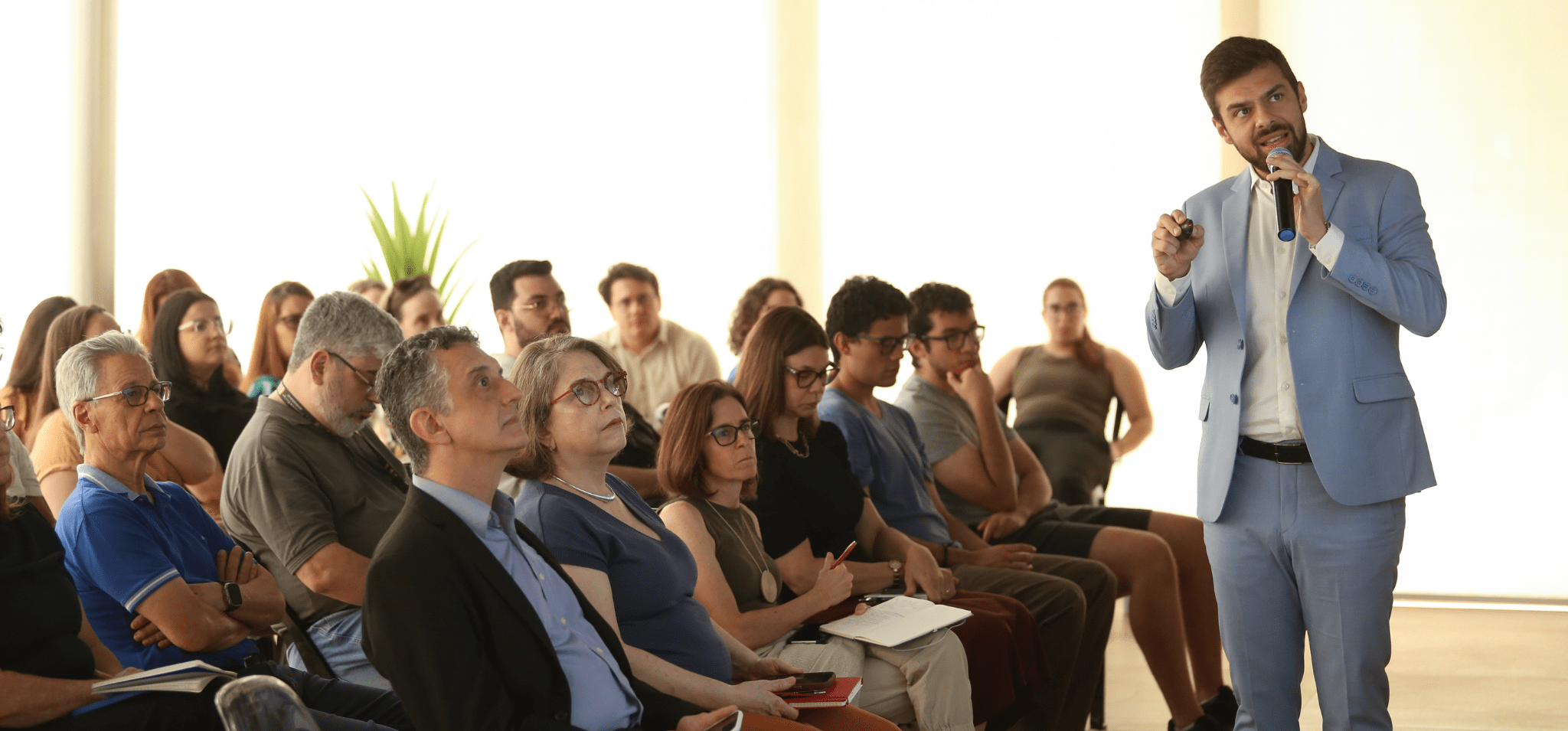 Uma fotografia colorida, em destaque no canto direiro está Rafael Carvalho de Fassio durante sua fala. Na esquerda, a platéia assiste atenta à apresentação. Fim da descrição.