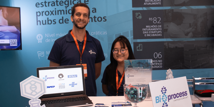 É possível ver na imagem duas pessoas posicionadas para a foto. Trata-se de um home e de uma mulher, ambos estão utilizando camisetas polo pretas. Eles estão centralizados em um stand que contém destaques do Parque Científico e Tecnológico da Unicamp ao fundo. Fim da descrição.