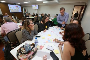 Foto colorida da sala Nova Iorque da Funcamp, onde aparecem os grupos sentados conversando entre si e escrevendo em papéis sobre mesas brancas. No primeiro plano, o professor Matheus conversa com um dos grupos. Fim da descrição.