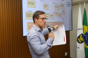 Foto colorida do professor Matheus Provinciali ministrando o workshop. Matheus é um homem branco, de cabelos curtos castanho claro. Ele usa camisa azul e óculos marrom. Na mão direita segura um microfone que posiciona embaixo da boca e na mão esquerda tem folhas brancas. Ao fundo aparecem parcialmente uma tela com projeção de texto e duas bandeiras, uma da cidade de Campinas e outra do Brasil. Fim da descrição.