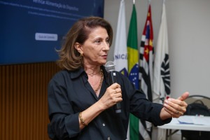 Foto colorida de Marilda, mulher branca de cabelos lisos castanho claro repicados na altura do ombro. Ela veste camisa preta, usa colares e pulseiras metátilcas e fala ao microfone. Ao fundo da sala estão posicionadas as bandeiras da cidade de Campinas, do Brasil, do estado de São Paulo e da Unicamp. Fim da descrição.