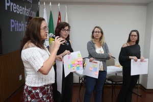 Foto colorida de apresentação de grupo formado por quatro mulheres brancas. Uma delas fala ao microfone e todas seguram na mão um cartaz com post-its coloridos colados. Ao fundo da sala lê-se no telão Presentacion Picth. Fim da descrição.