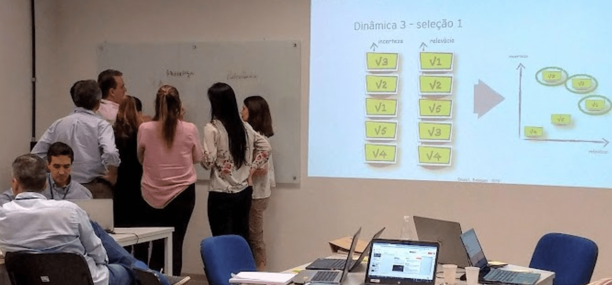 Foto de uma sala de aula com várias pessoas. No primeiro plano, há mesas organizadas com notebooks e materiais de anotação. Ao fundo, um grupo de pessoas está reunido em frente a um quadro branco, discutindo um tema projetado no telão. O ambiente tem paredes brancas, cadeiras azuis e iluminação artificial. Os participantes estão concentrados na atividade. Fim de descrição.