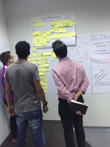 Foto de três homens em pé diante de um painel branco coberto por post-its amarelos. Um deles segura um caderno preto enquanto observa as anotações. O ambiente tem paredes brancas e outros papéis afixados com informações escritas à mão. O grupo parece estar analisando estratégias ou mapeando um processo. Fim de descrição.