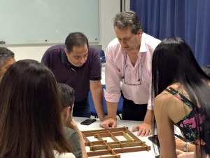 Foto de um grupo de pessoas reunidas ao redor de uma mesa. No centro da imagem, dois homens observam um tabuleiro de madeira com peças organizadas em um padrão. Outras pessoas ao redor também acompanham a atividade. O ambiente tem cortinas azuis ao fundo e iluminação artificial. Todos demonstram concentração e engajamento. Fim de descrição.