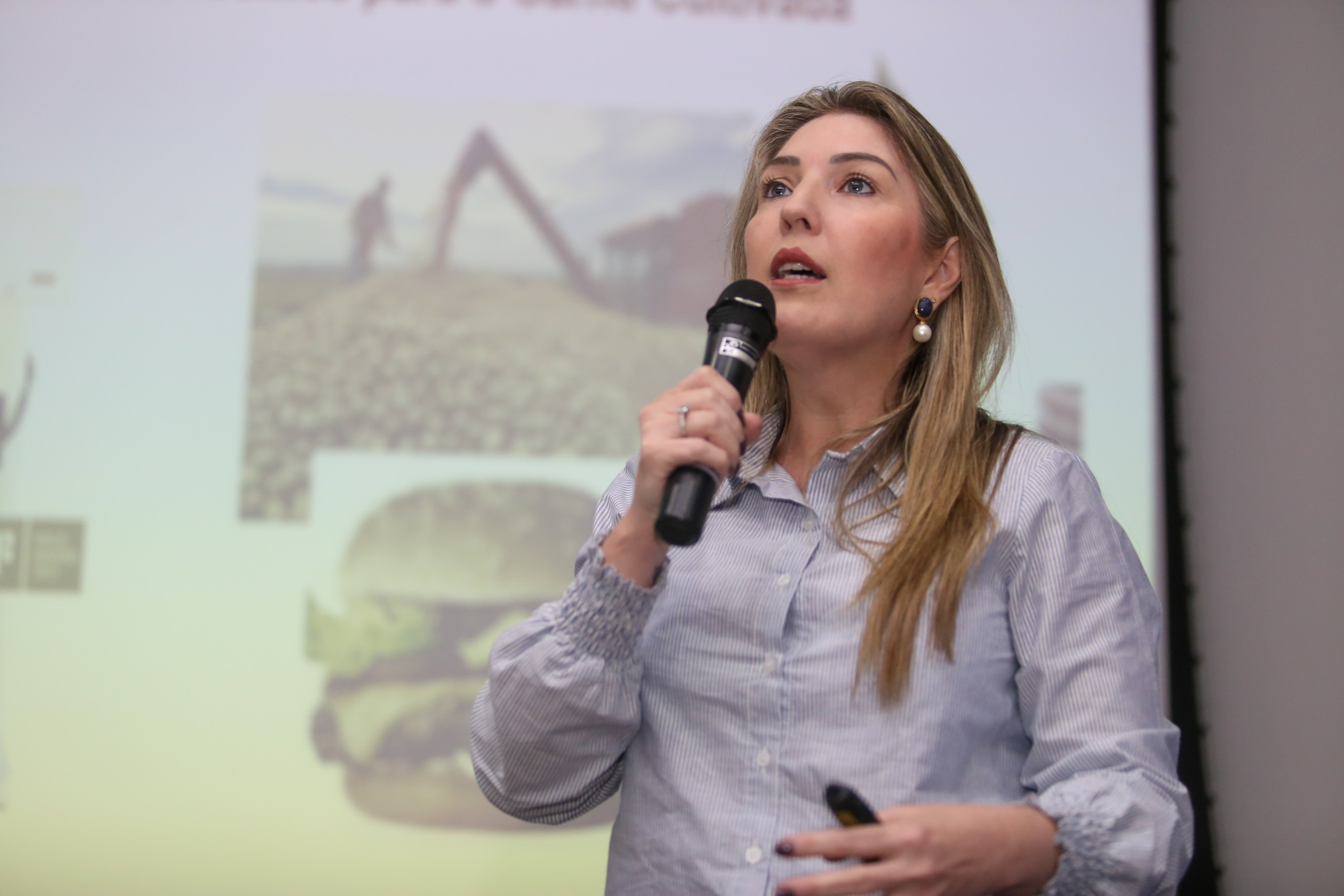 Foto de uma mulher falando ao microfone e olhando para a frente, onde está a plateia, que fica oculta na imagem. Ela é uma mulher loira, de cabelos longos e usa uma camisa social. Fim da descrição. 