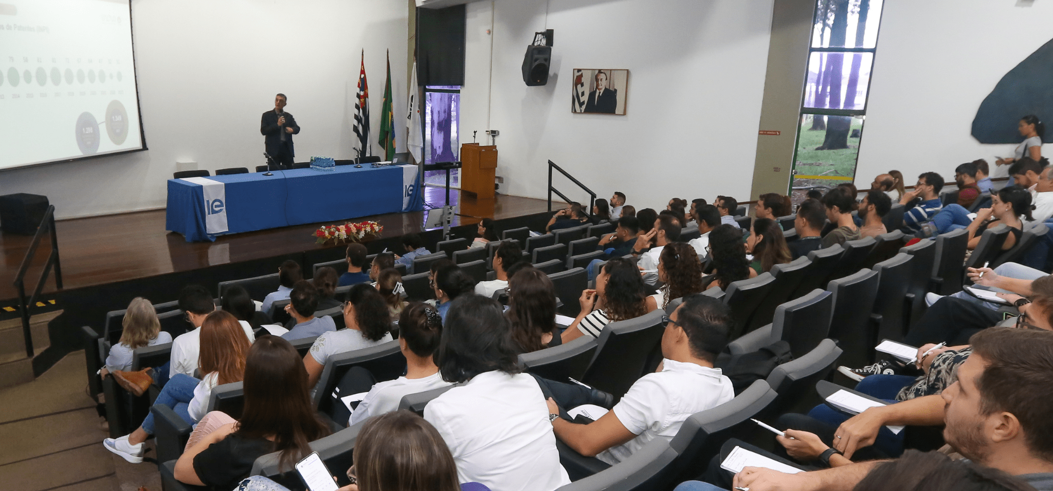 Foto de plateia durante o evento de inovação em biotecnologia. Na imagem, feita em um auditório fechado, há diversas pessoas de costas para foto e olhando para o palestrante, que está de frente para a câmera. Fim da descrição.