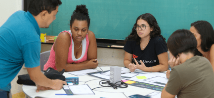 Na foto estão quatro mulheres sentadas envolta de uma mesa redonda prestando atenção na explicação do Vital, um homem, branco, com cabelos pretos. Ele está explicando sobre como montar um modelo de negócios usando o Canvas. A oficina aconteceu na Semana do Empreendedorismo de 2024. Fim da descrição.