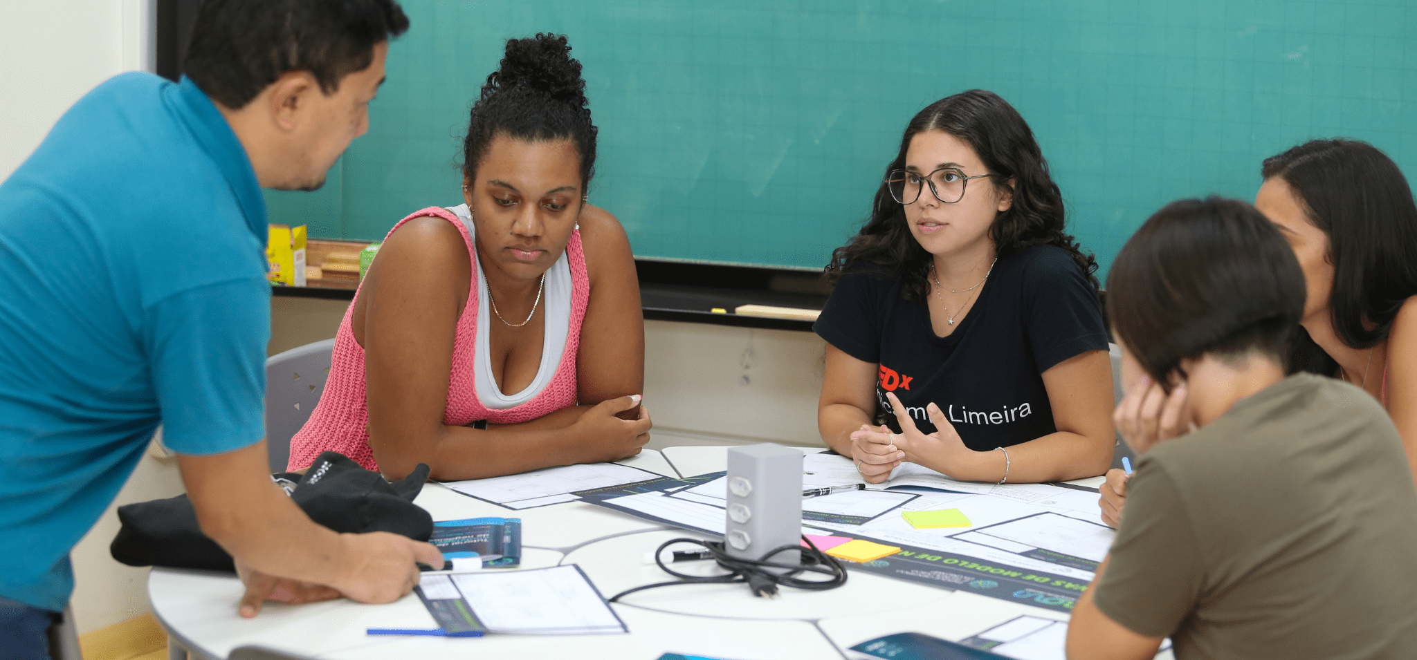 Na foto estão quatro mulheres sentadas envolta de uma mesa redonda prestando atenção na explicação do Vital, um homem, branco, com cabelos pretos. Ele está explicando sobre como montar um modelo de negócios usando o Canvas. A oficina aconteceu na Semana do Empreendedorismo de 2024. Fim da descrição.